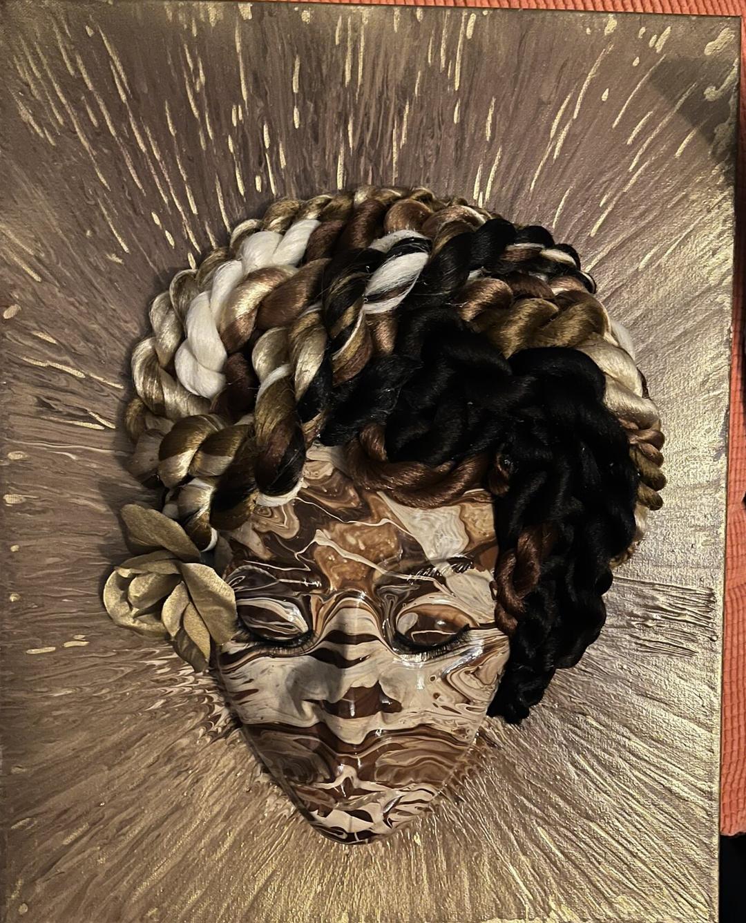 A sculpted face with marbled brown and cream paint, framed by twisted black, brown and gold braids and gold leaves, on a gold splattered canvas.