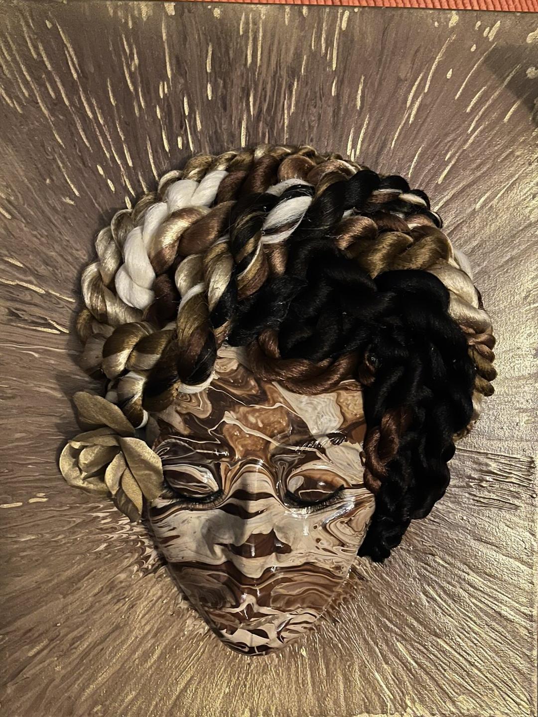 A sculpted face with marbled brown and cream paint, framed by twisted black, brown and gold braids and gold leaves, on a gold splattered canvas.